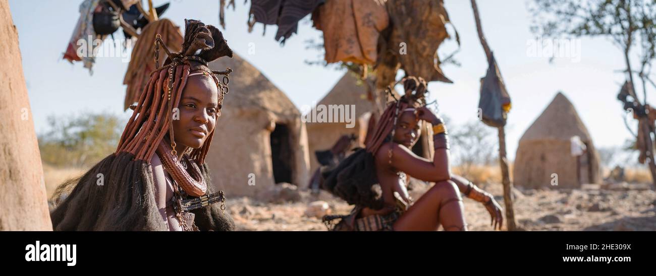 Panoramaaufnahme mit Himba-Frauen, die vor ihren Hütten in einem traditionellen Himba-Dorf in der Nähe von Kamanjab im Norden Namibias, Afrika, sitzen. Stockfoto