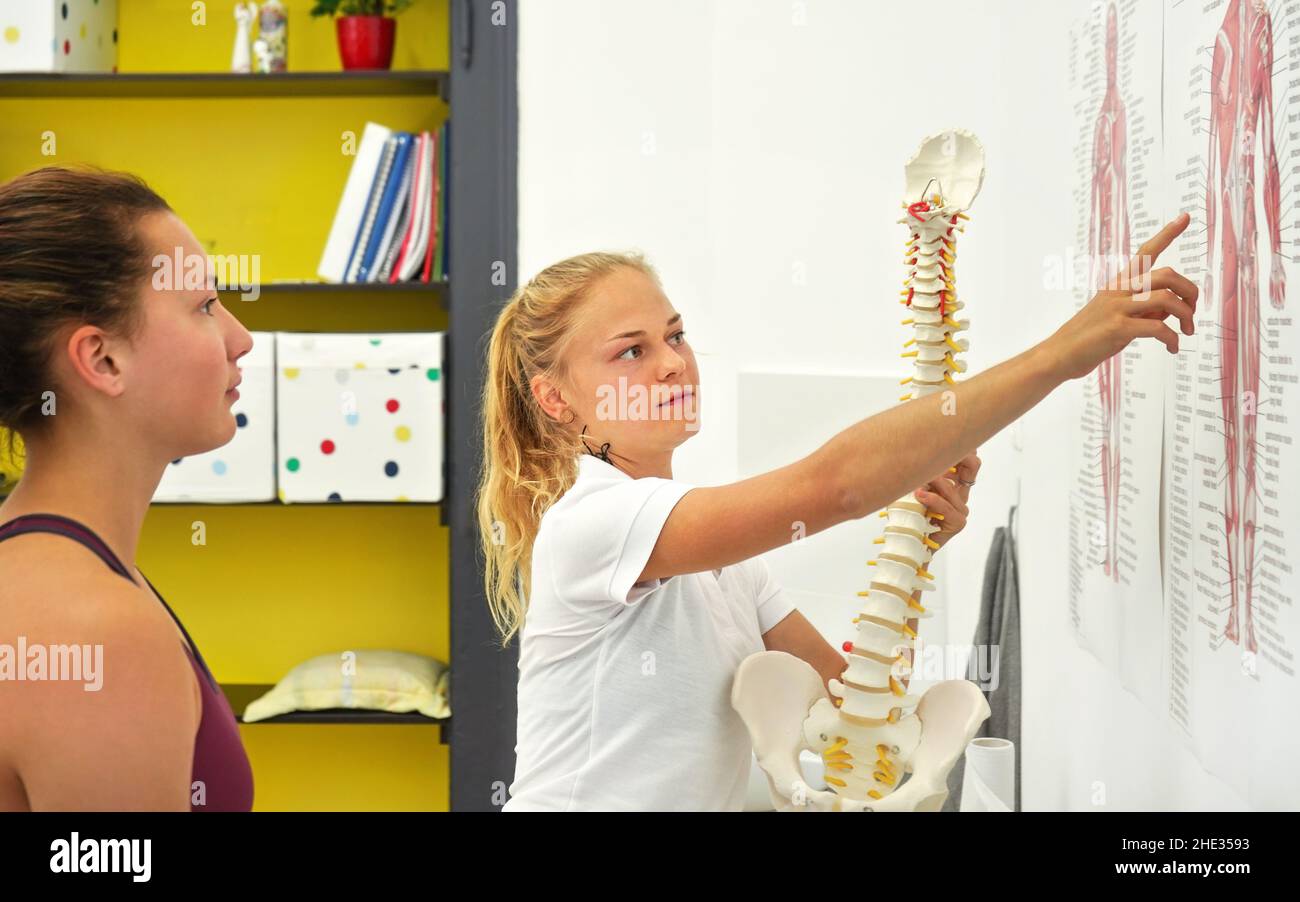 Junge Frau Patientin bei der Physiotherapie-Beratung, Physiotherapeutin erklärt Rückenknochen auf menschliche Muskeln Körperdiagramm, hält Wirbelsäulenmodell Stockfoto