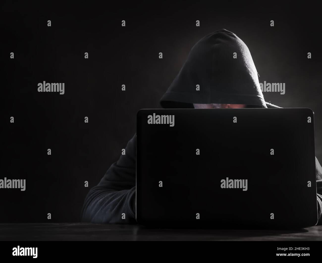 Hinter dem Laptop sitzender Hacker mit Kapuze vor dunklem Hintergrund Stockfoto