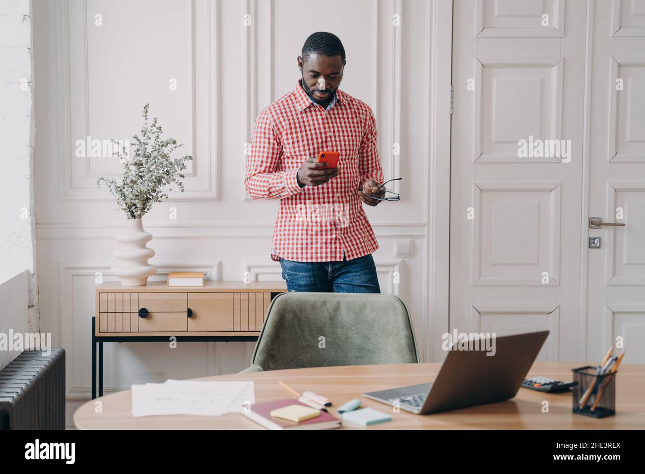 Junger seriöser afroamerikanischer Geschäftsmann in legerer Kleidung, der mit dem Smartphone im Büro steht Stockfoto