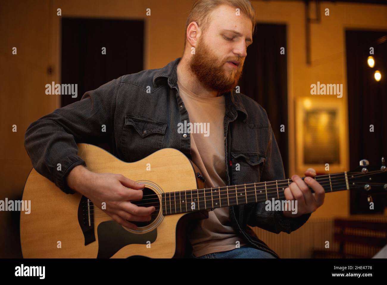 Ein junger Mann mit Bart spielt in einem Raum mit warmer Beleuchtung eine akustische Gitarre Stockfoto