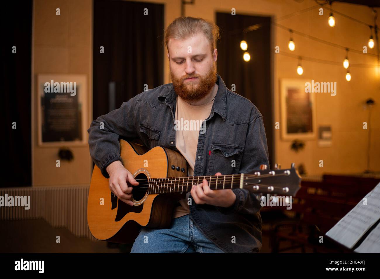 Ein junger Mann mit Bart spielt in einem Raum mit warmer Beleuchtung eine akustische Gitarre Stockfoto