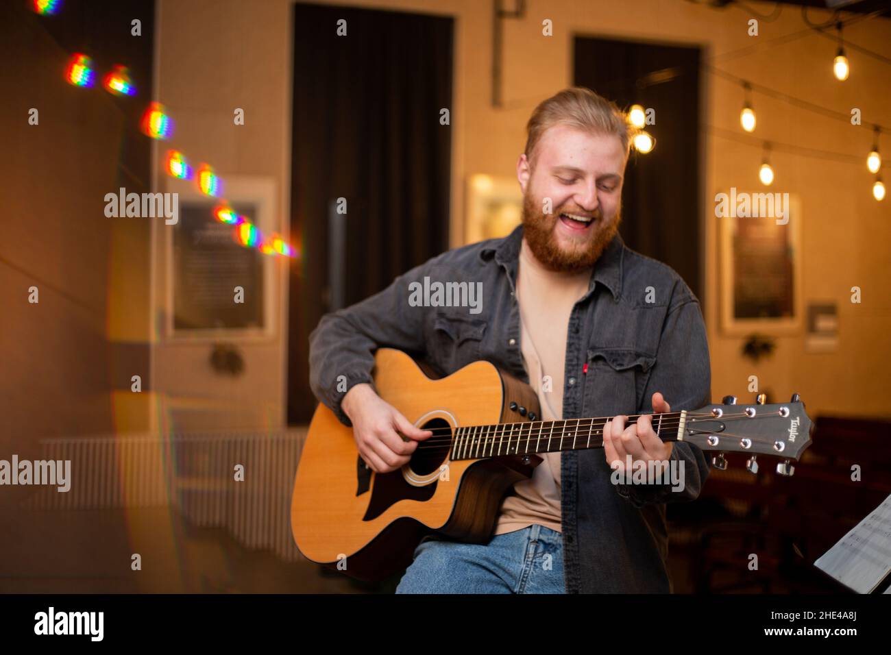 Ein junger Mann mit Bart spielt in einem Raum mit warmer Beleuchtung eine akustische Gitarre Stockfoto