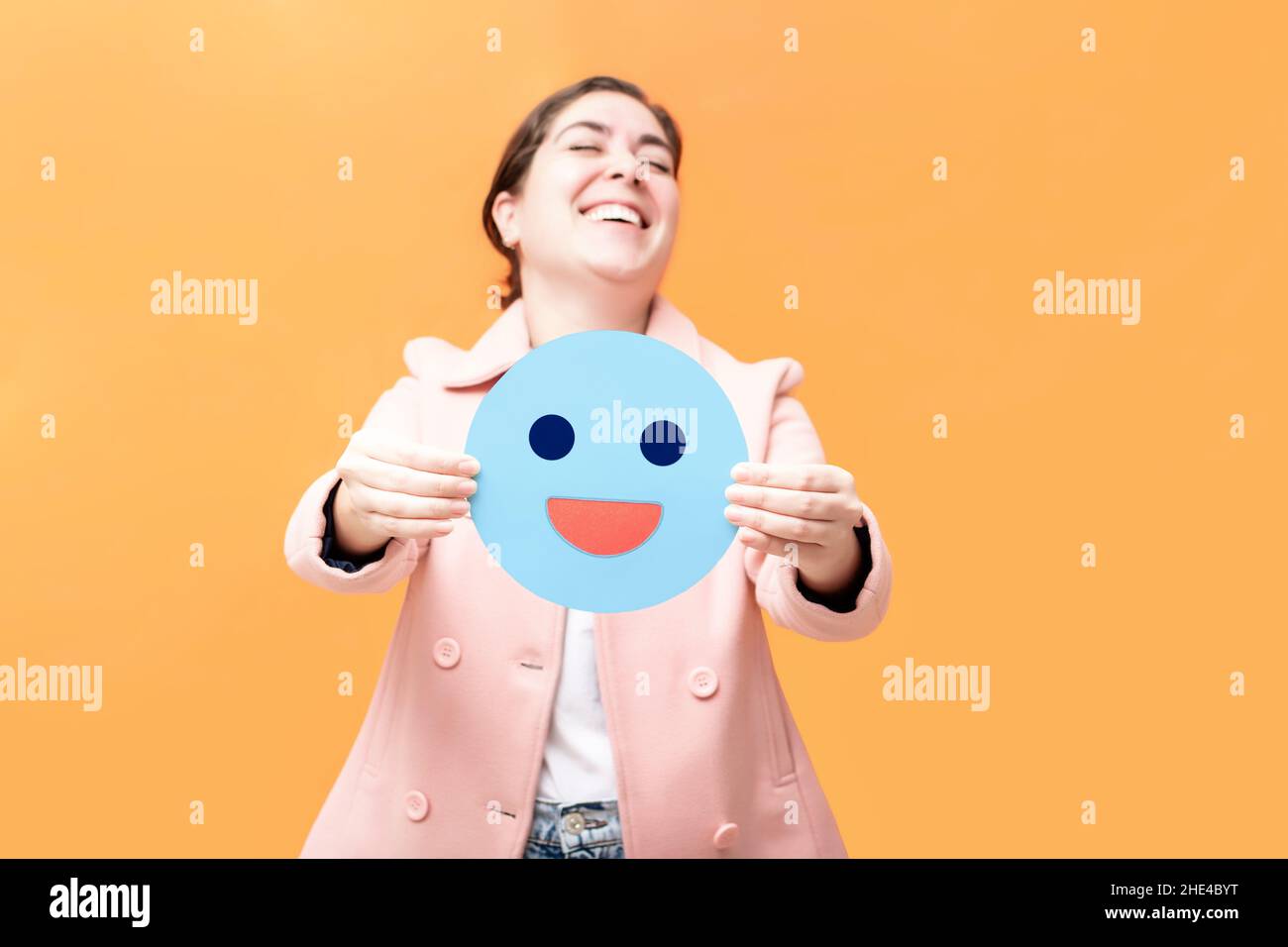 Glückliches Mädchen hält ein Stück Papier mit blauen Emoji, isoliert auf orangefarbenem Hintergrund. Blue monday-Konzept. Stockfoto