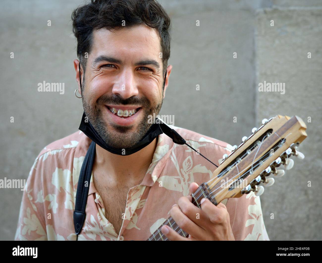 Ein hübscher junger Lateinamerikaner mit schwarzem Bart und Gesichtsmaske unter dem Kinn spielt eine kleine Andengitarre (Charango) mit zehn Saiten. Stockfoto