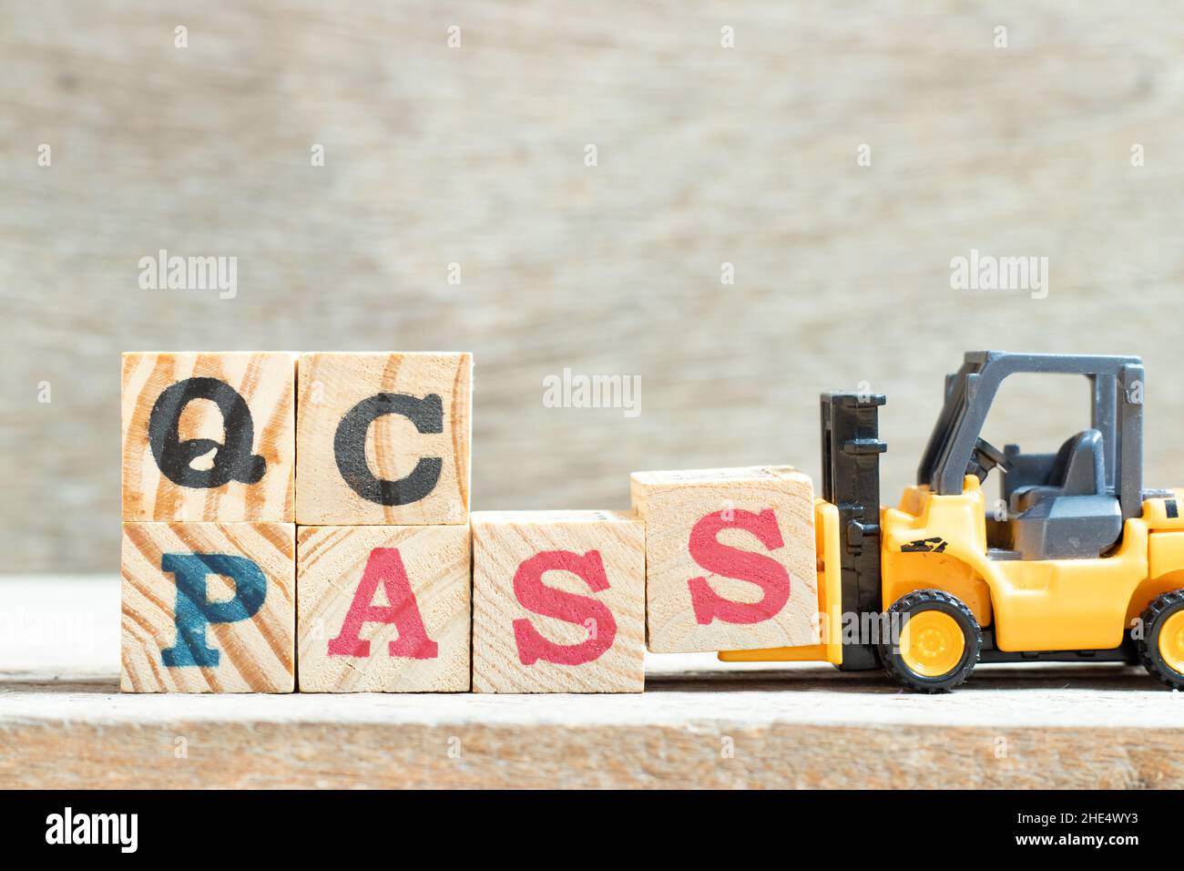 Spielzeug-Gabelstapler halten Buchstabenblock s, um Wort QC (Qualitätskontrolle) Passon Holzhintergrund abzuschließen Stockfoto