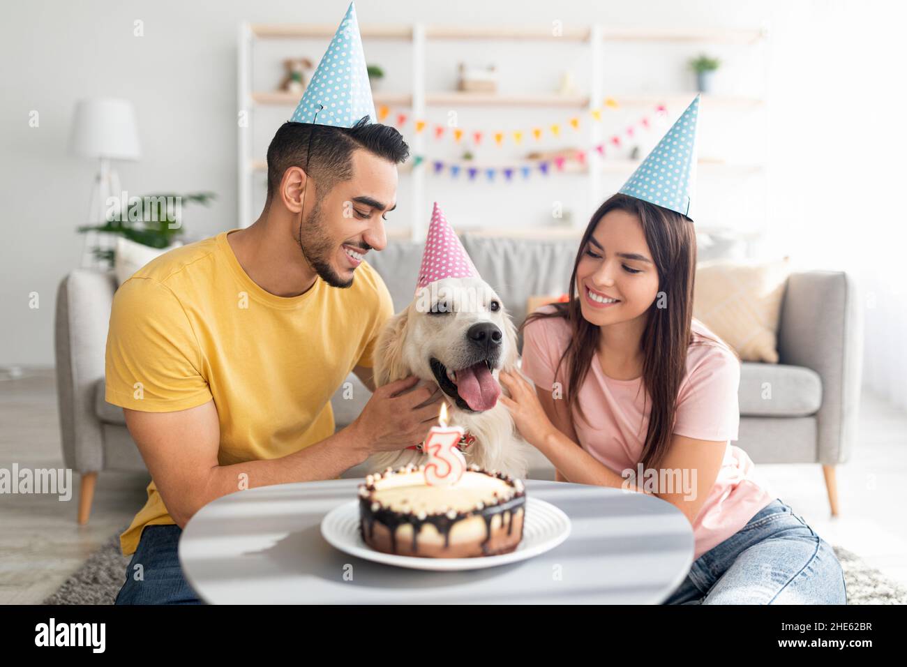 Fröhliche, vielfältige Ehepartner feiern den Geburtstag ihres Hundes mit einem leckeren Kuchen und tragen zu Hause festliche Hüte. Feiertagsfeier Stockfoto
