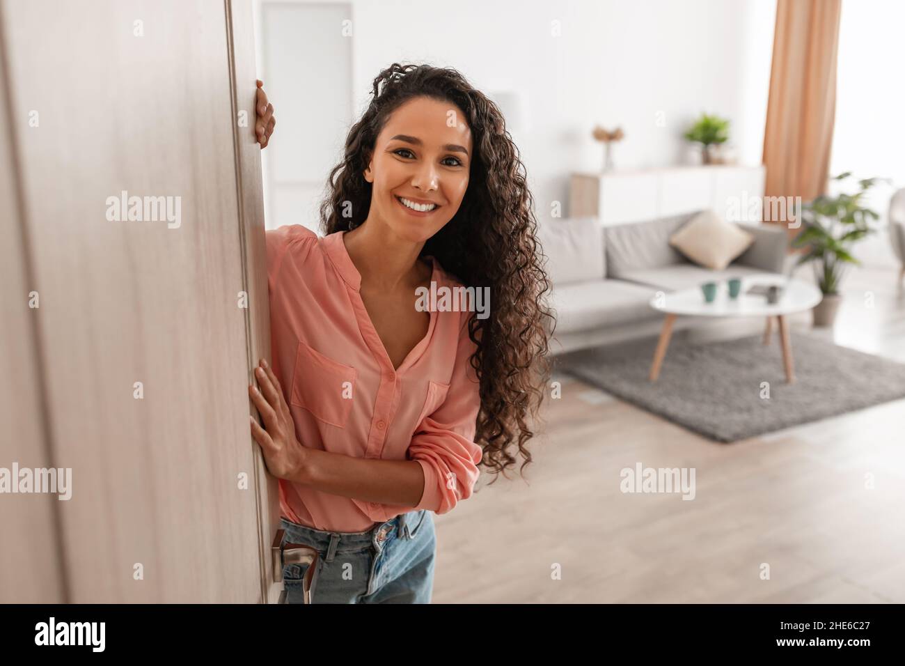 Fröhliche junge lockige Dame, die Leute zum Betreten des Hauses einlädt Stockfoto