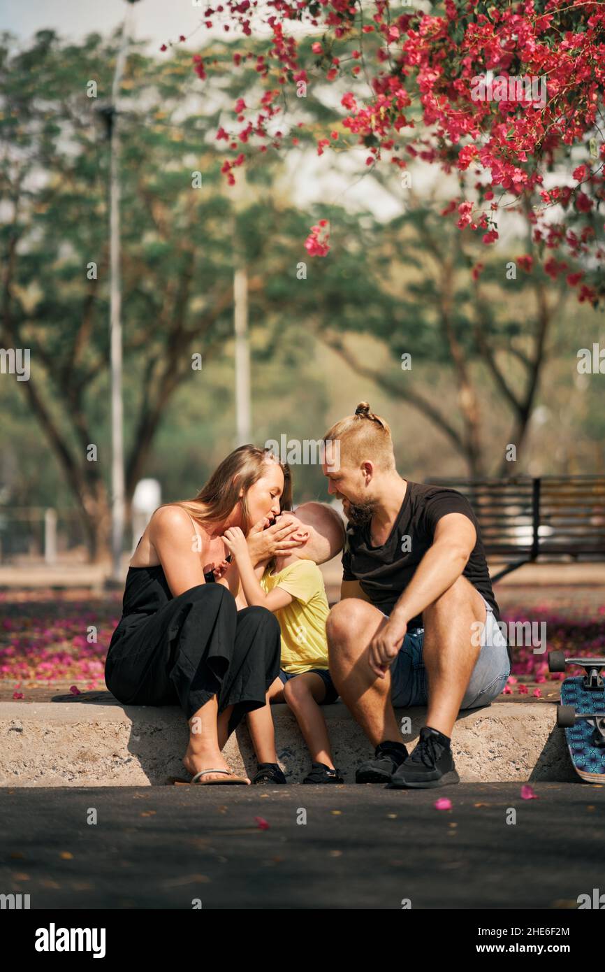 Vater und Mutter umarmen ihren kleinen Sohn, während sie im Skatepark mit Blütenbäumen herumsitzen. Konzept der Liebe und Fürsorge der Familie. Freizeit im Freien Stockfoto