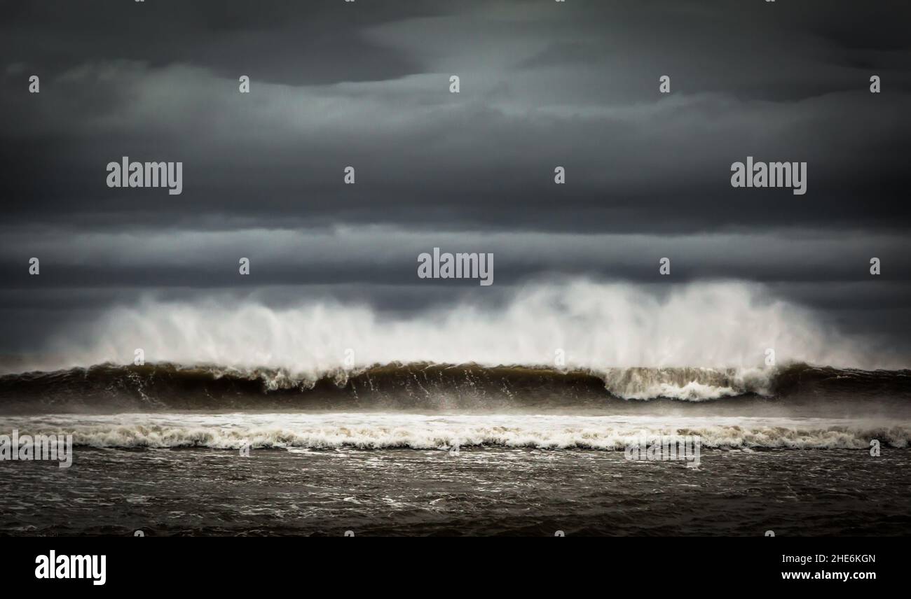 Eine große Welle beginnt zu brechen, während sie sich dem Ufer nähert, während die Windstöße von den Spitzen sprühen Stockfoto