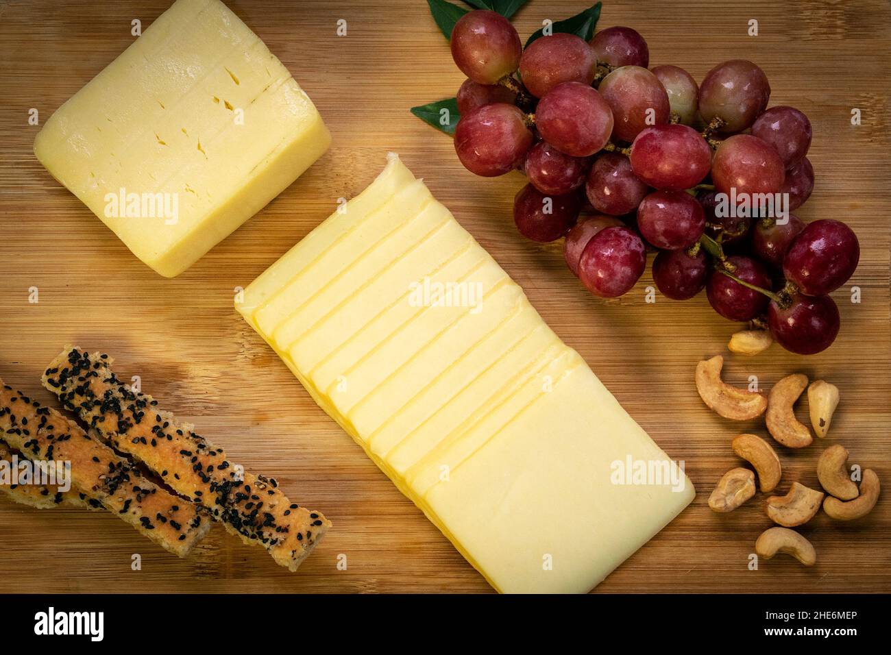 Cheddar-Käseblock in Scheiben auf Holzbrett mit Trauben, Cashewnüssen, Brotstäbchen und Honig Stockfoto