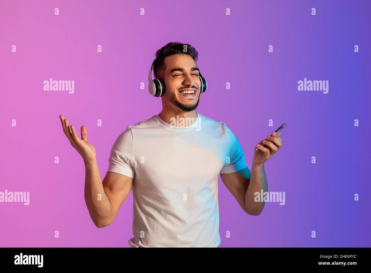 Cooler arabischer Mann, der in Kopfhörern beliebte Lieder hört, die neue mobile Musik-App am Telefon überprüft und sich im Neonlicht entspannt Stockfoto