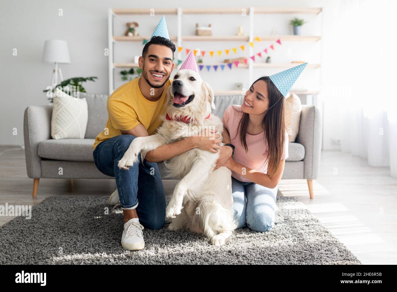 Ein junges, multinationales Paar in Partyhüten feiert den Geburtstag des Hundes, posiert und lächelt vor der Kamera Stockfoto
