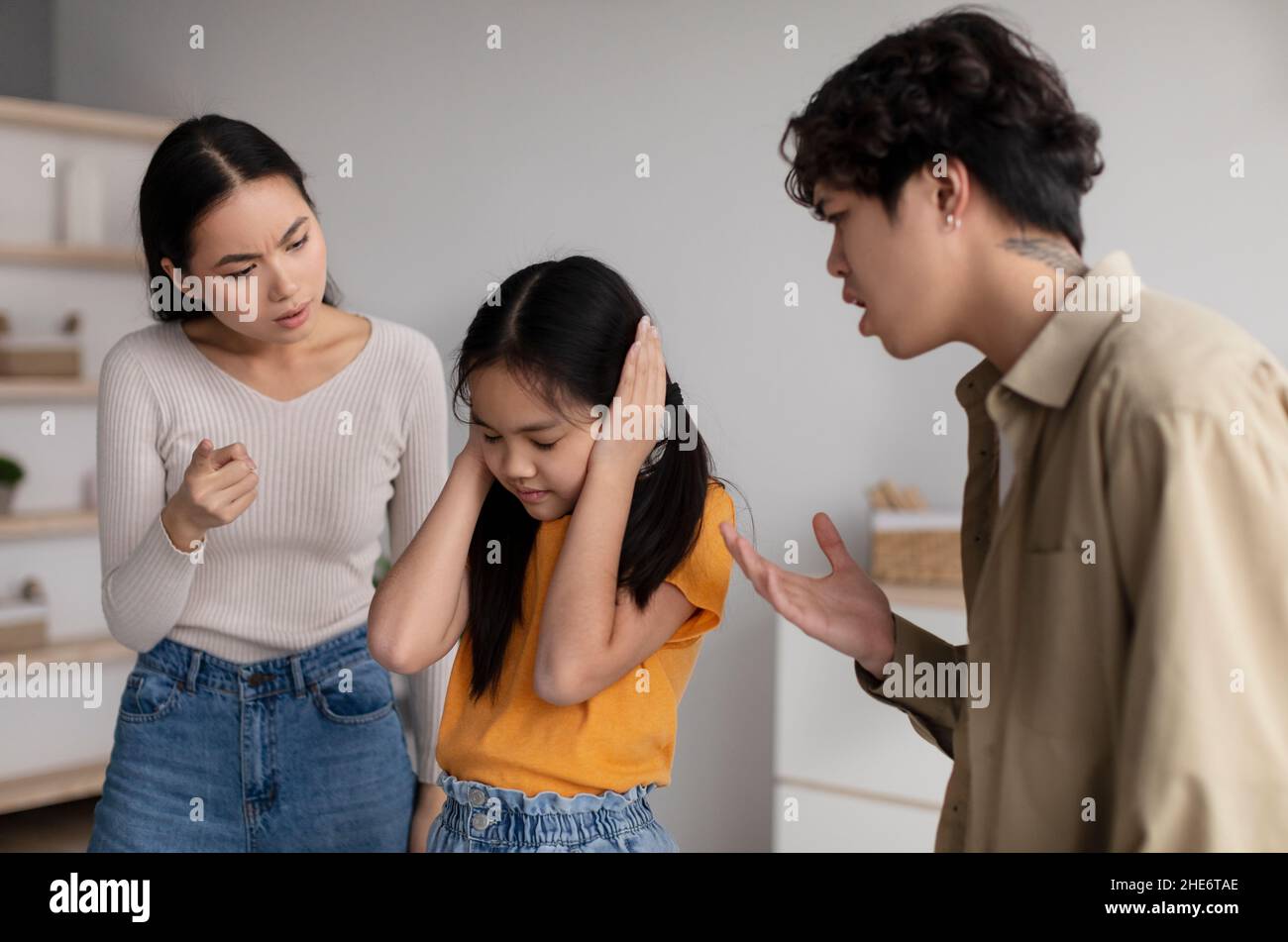 Verzweifelte kleine Teenager-Tochter schließt ihre Ohren, um ihre Eltern, ihre junge asiatische Dame und ihren Kerl nicht zu hören, der Mädchen schimpft Stockfoto
