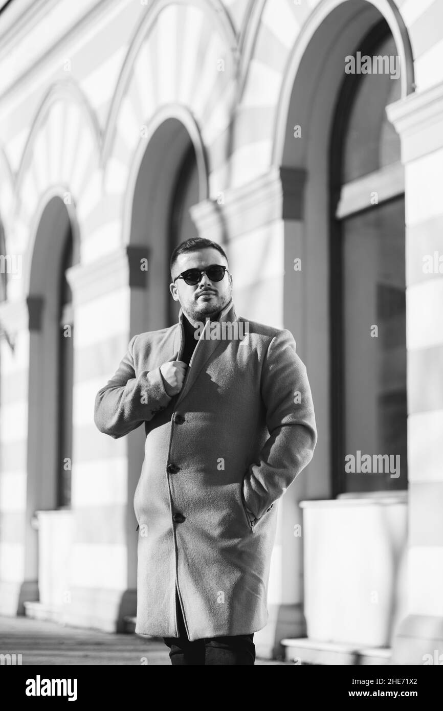 Ein kaukasischer Mann in modischem Mantel und Sonnenbrille, der in der Nähe eines schönen Gebäudes steht Stockfoto