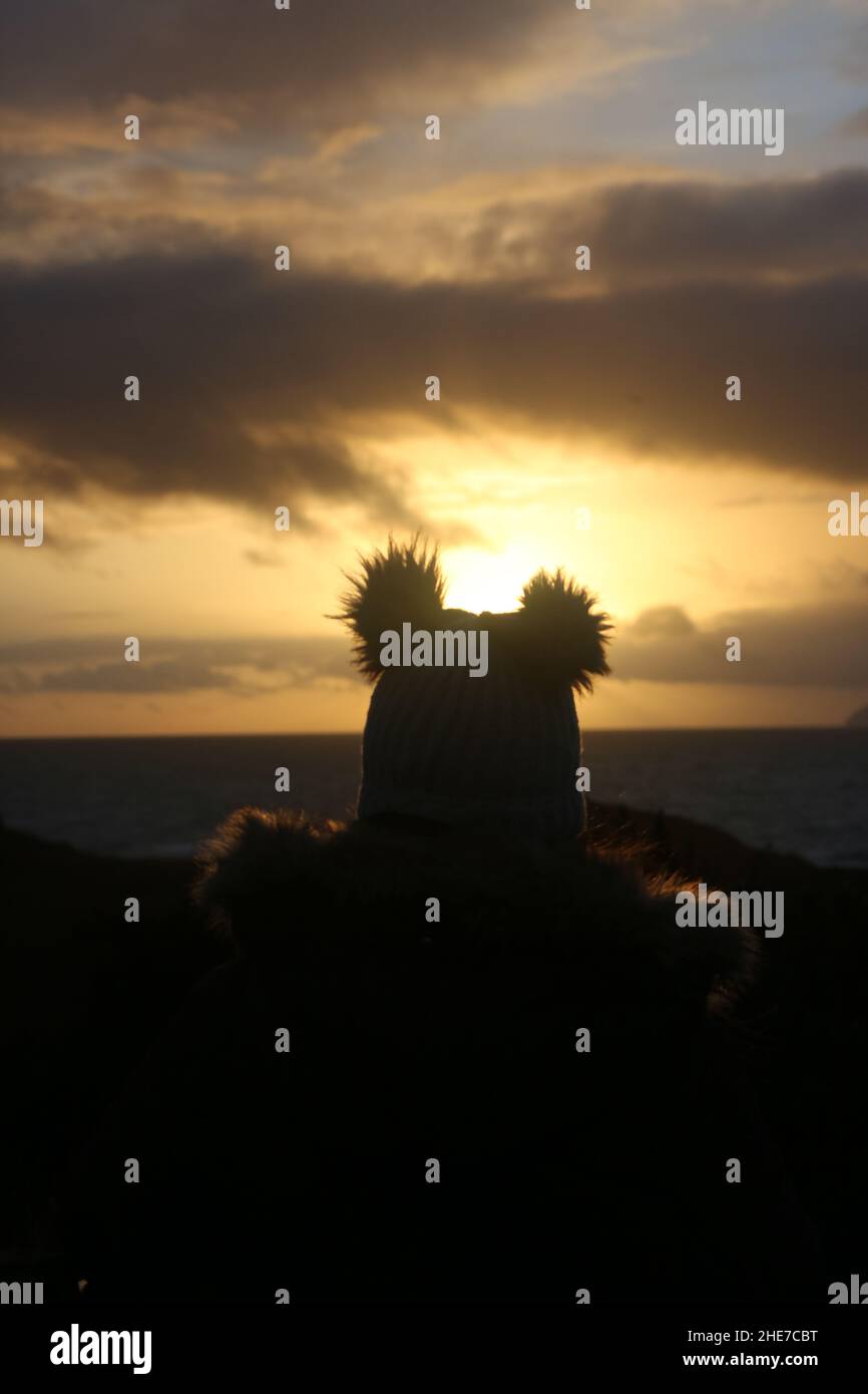 Dunure, Ayrshire, Schottland, Großbritannien. Eine Frau am Wintertag sitzt mit dem Rücken zur Kamera und schaut den Sonnenuntergang an der Ayrshire Coast an, mit einem warmen Bommel-Hut Stockfoto