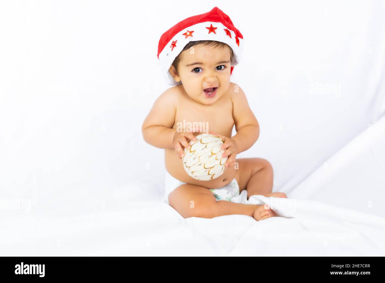 Ein kleiner Junge mit einem roten Weihnachtshut auf weißem Hintergrund, Copy Space, mit Weihnachtsmann-Hut, der mit einem Weihnachtsbaumball spielt Stockfoto