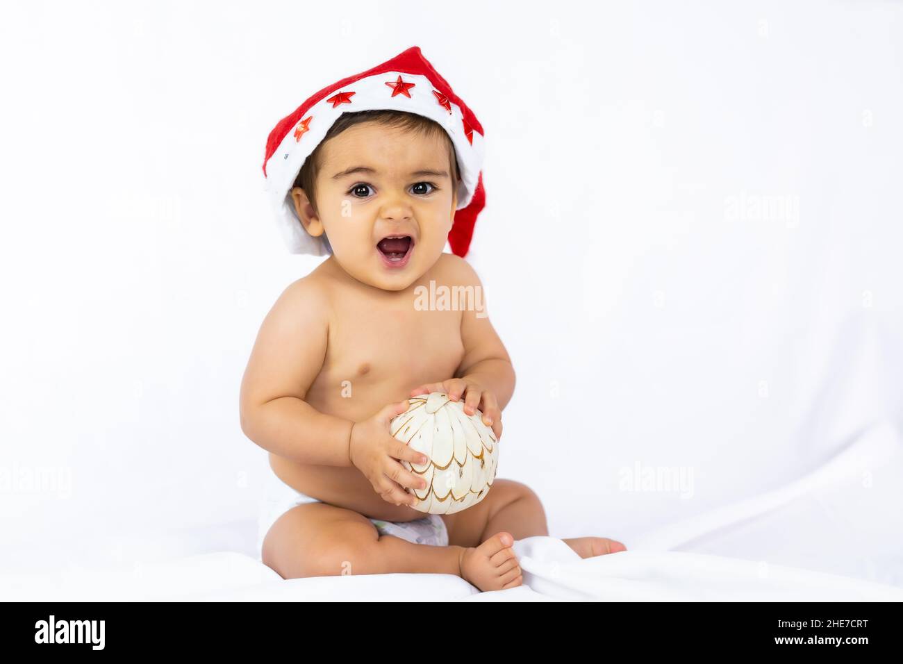Ein kleiner Junge mit einem roten Weihnachtshut auf weißem Hintergrund, Copy Space, mit Weihnachtsmann-Hut, der mit einem Weihnachtsbaumball spielt Stockfoto