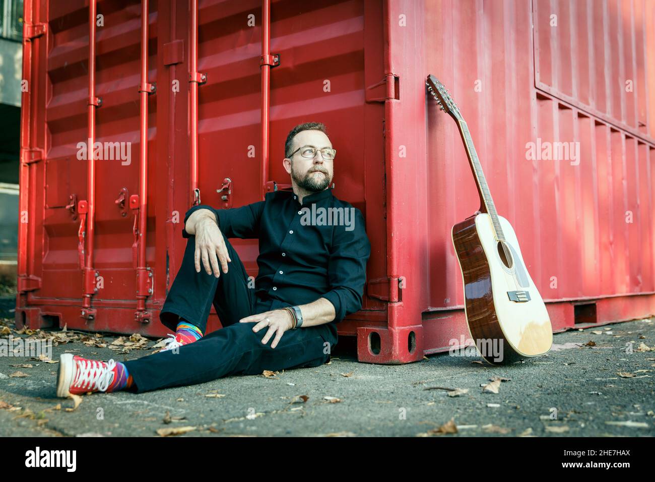 Mann in Schwarz mit einem Bart, der auf einem Boden unter einem roten Behälter mit einer akustischen 12-saitigen Gitarre sitzt Stockfoto