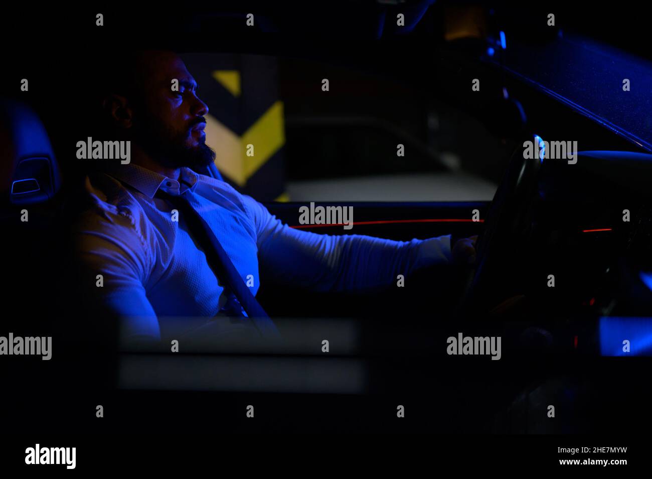 Niedlicher bärtiger, starker junger Mann in weißem Hemd mit Krawatte sitzt in einem luxuriösen Sport-Muskelwagen, der mit blauem Licht in einer Tiefgarage beleuchtet ist Stockfoto