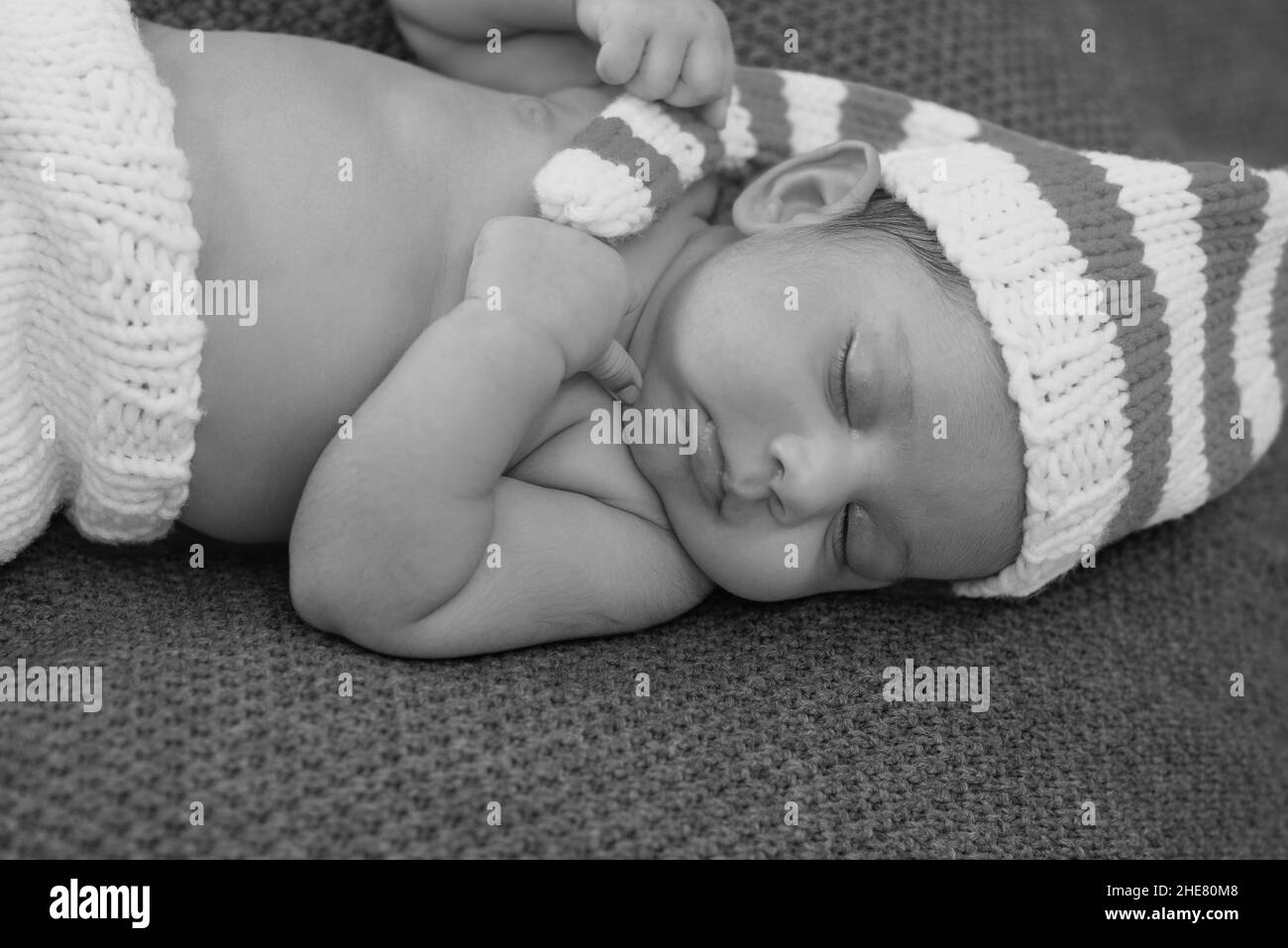 Die Graustufenaufnahme eines liegenden Neugeborenen in einem gestreiften Hut schläft mit zusammengeklemmten Händen Stockfoto