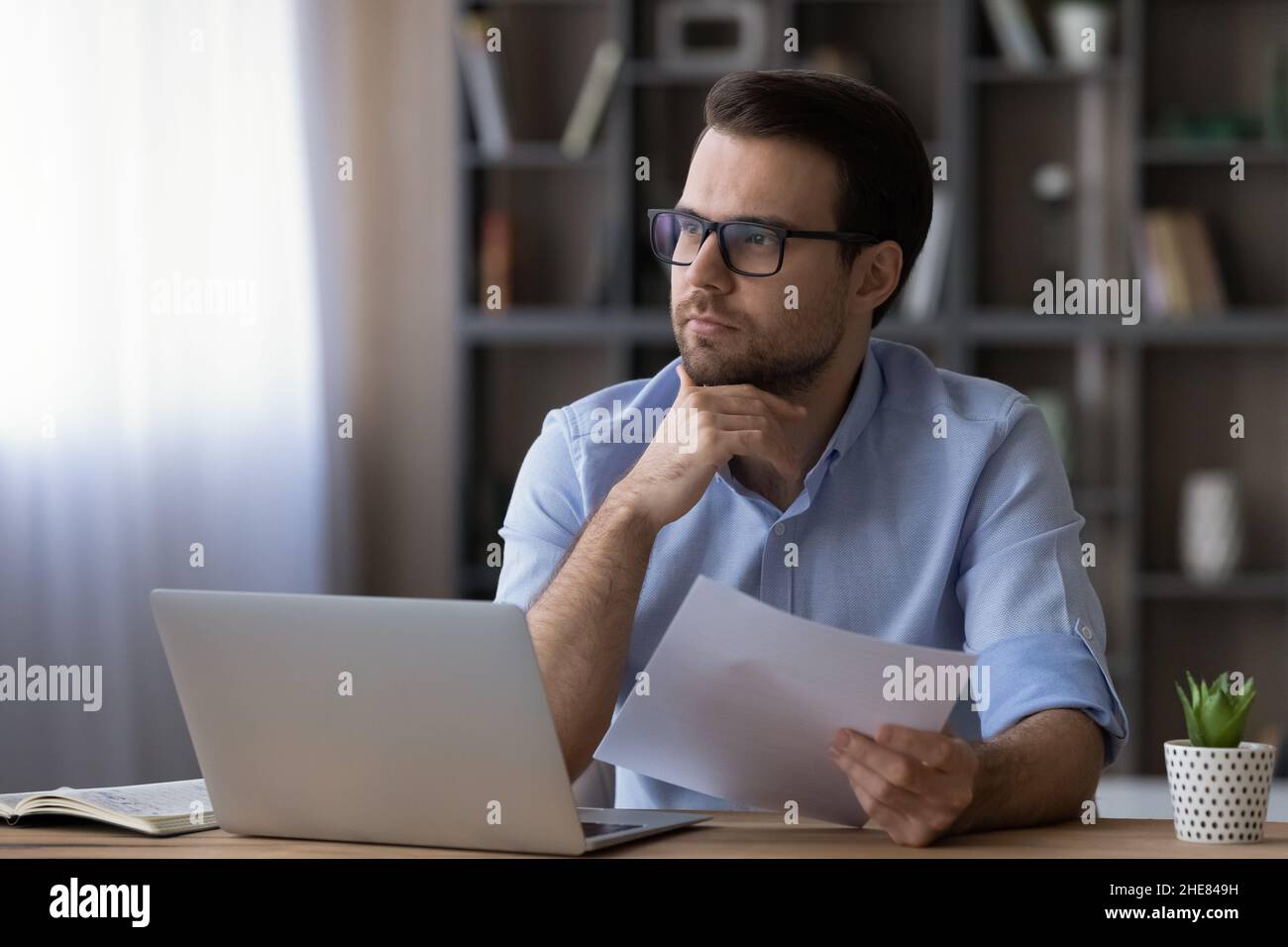 Nachdenklicher ernsthafter Geschäftsmann in einer Brille, der das Kinn berührt und das Dokument hält Stockfoto