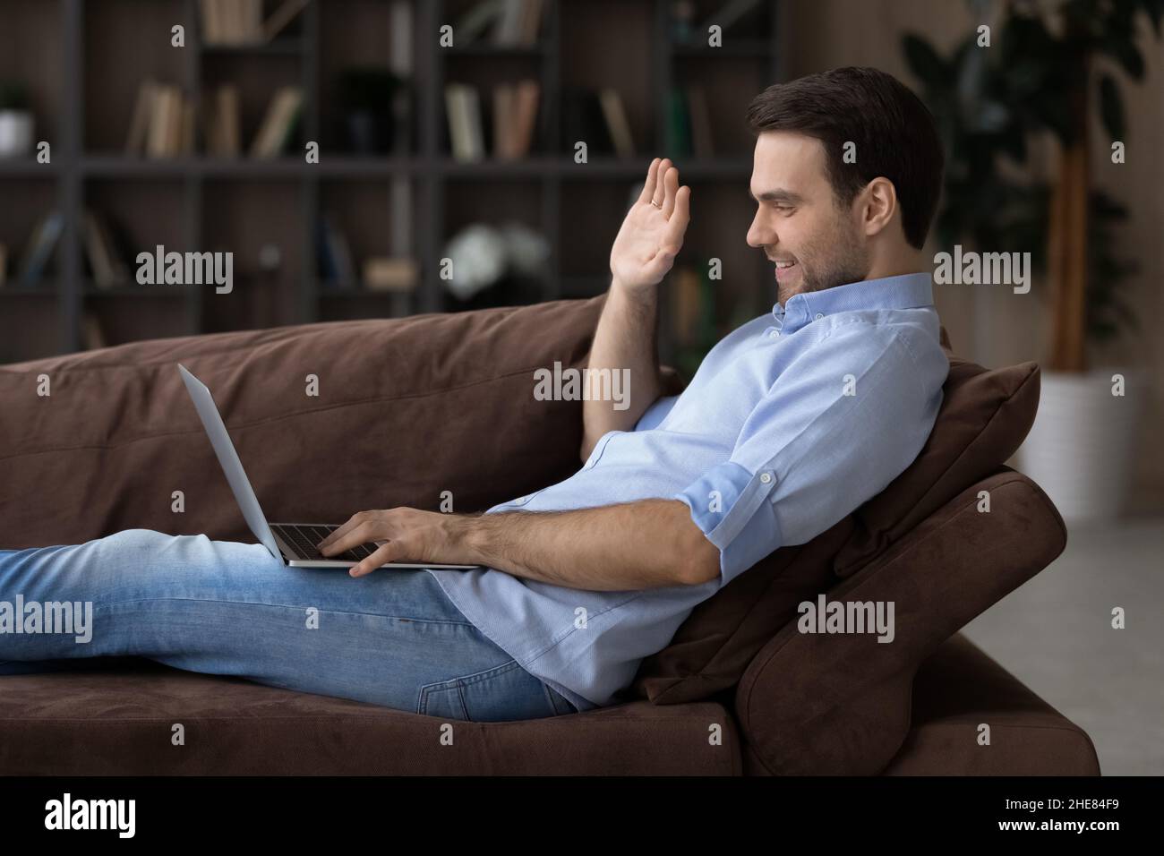 Profilansicht lächelnder Mann winkt mit der Hand und benutzt den Laptop auf der Couch Stockfoto