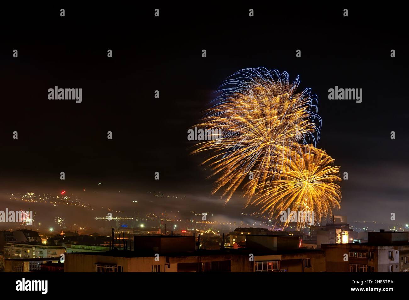 Leuchtend goldblaues Feuerwerk funkelt am dunklen Himmel über der Stadt. Die kleine Stadt feiert das neue Jahr mit bunten Pyrotechnik in der Nacht. Feiertagsfeuerwerk. Stockfoto