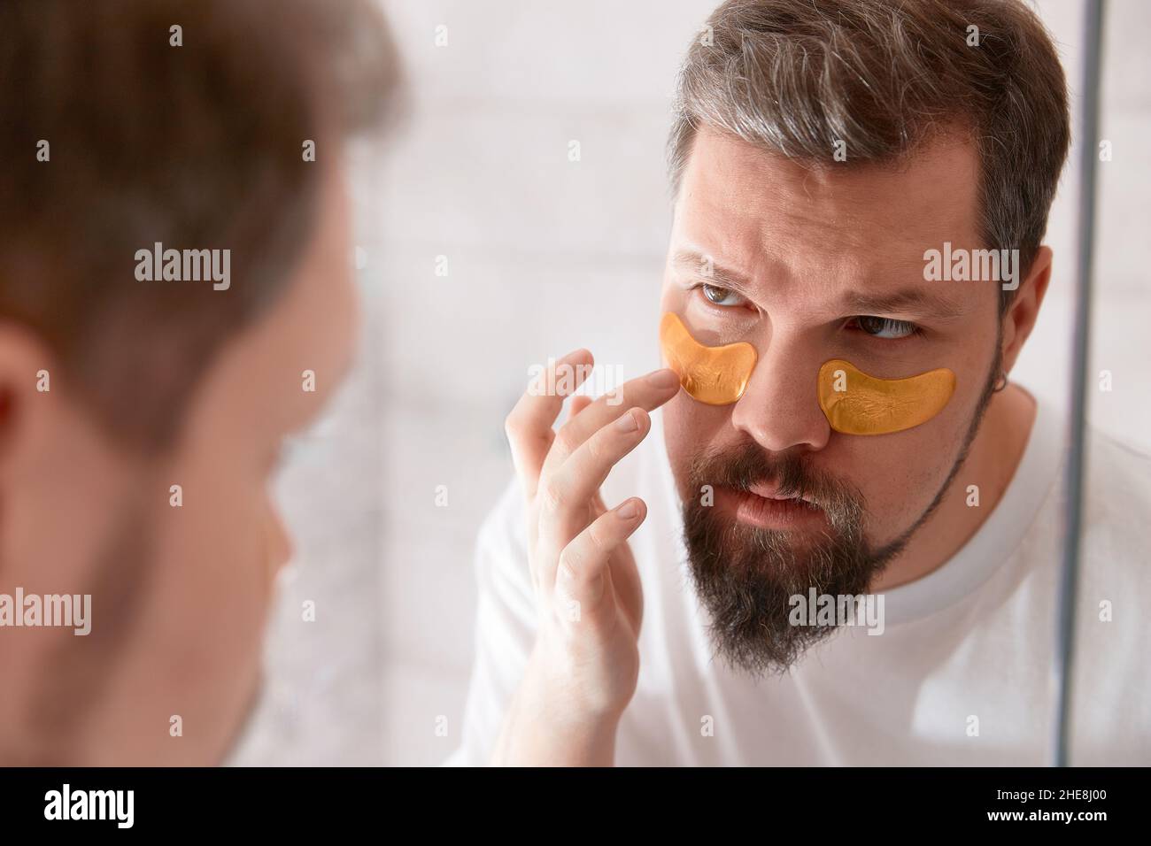 Porträt eines Mannes in weißem T-Shirt, der im Badezimmer Augenflecken auf sein Gesicht aufträgt. Self-Care-Verfahren am Morgen Stockfoto