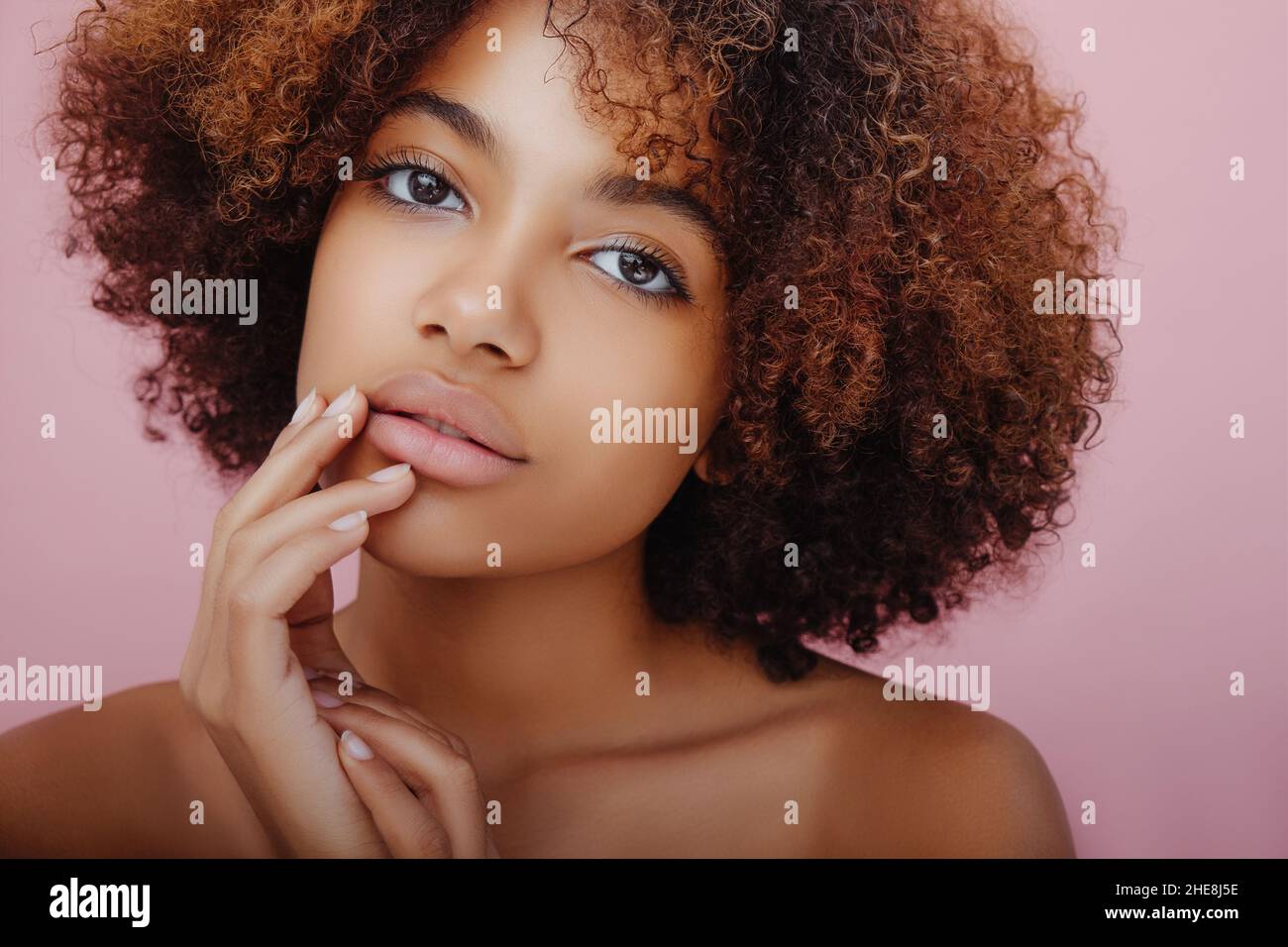 Schönheitsporträt einer schönen afroamerikanischen jungen Frau mit lockigem Haar, die geheimnisvoll auf die Kamera schaut und ihre Finger auf einem rosa Hintergrund auf ihr Gesicht legt Stockfoto