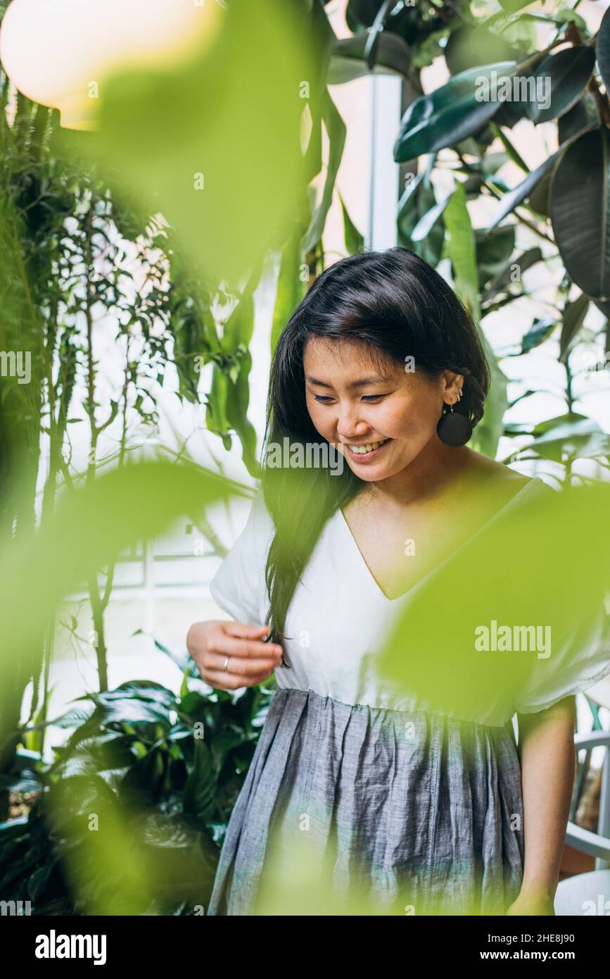 Junge Asiatin mit langen schwarzen Haaren in weiß-grauem Kleid sieht glücklich aus und lächelt unter grünen Pflanzen in veganer Café-Nahaufnahme Stockfoto