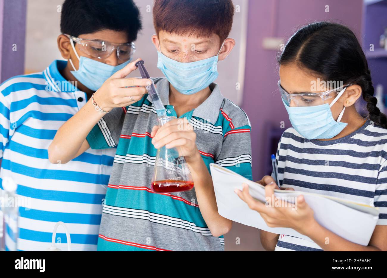 Diverse Kinder experimentieren mit Sicherheitsbrillen und Gesichtsmaske, indem sie Ergebnisse beim Mischen von Chemikalien mit Kolben im Chemisterielabor notieren - Konzept Stockfoto