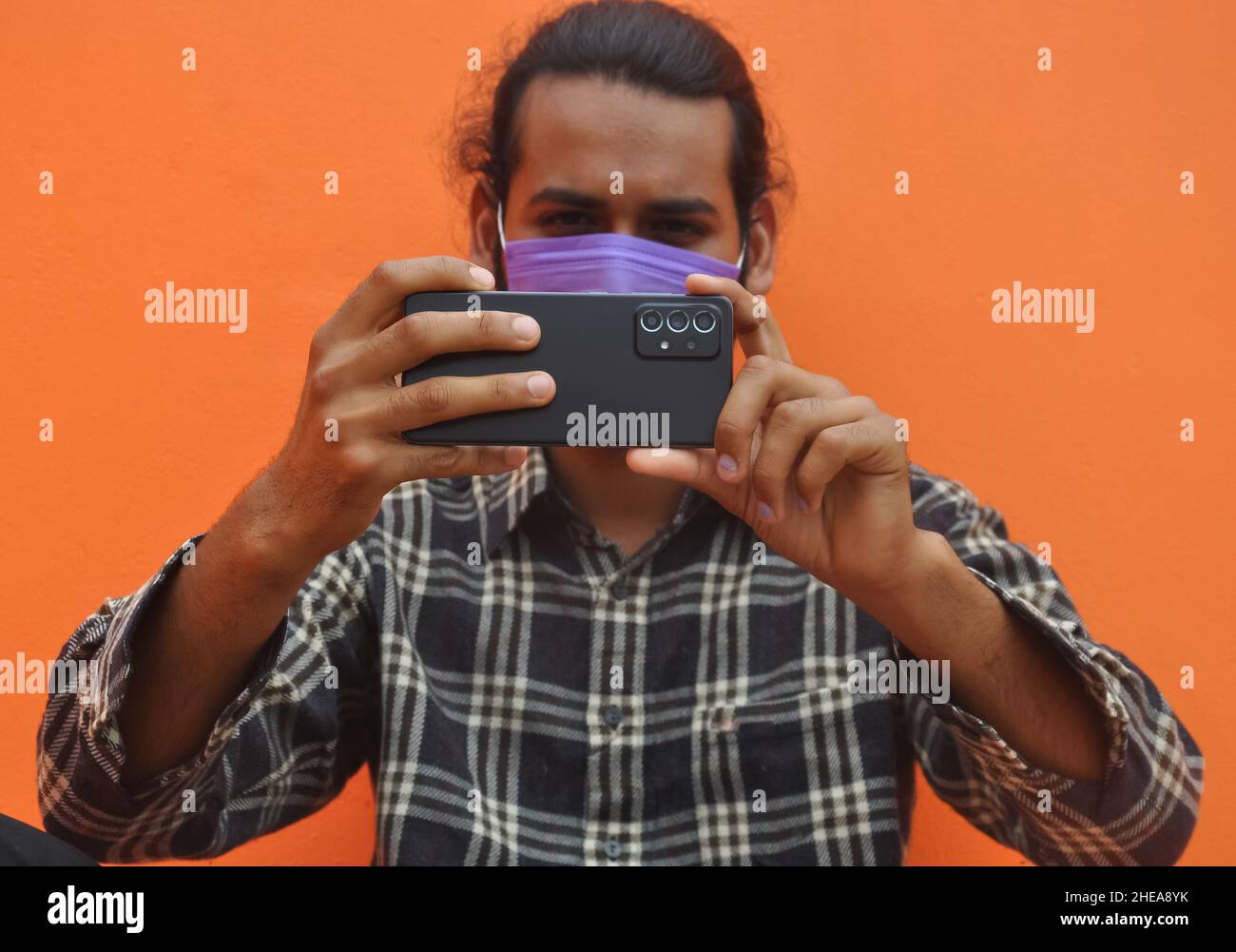 Selektiver Fokus eines jungen Mannes, der eine Gesichtsmaske trägt und Sie mit seinem Mobiltelefon fotografiert, sich gegen eine orangefarbene Wand mit Kopierraum posiert Stockfoto