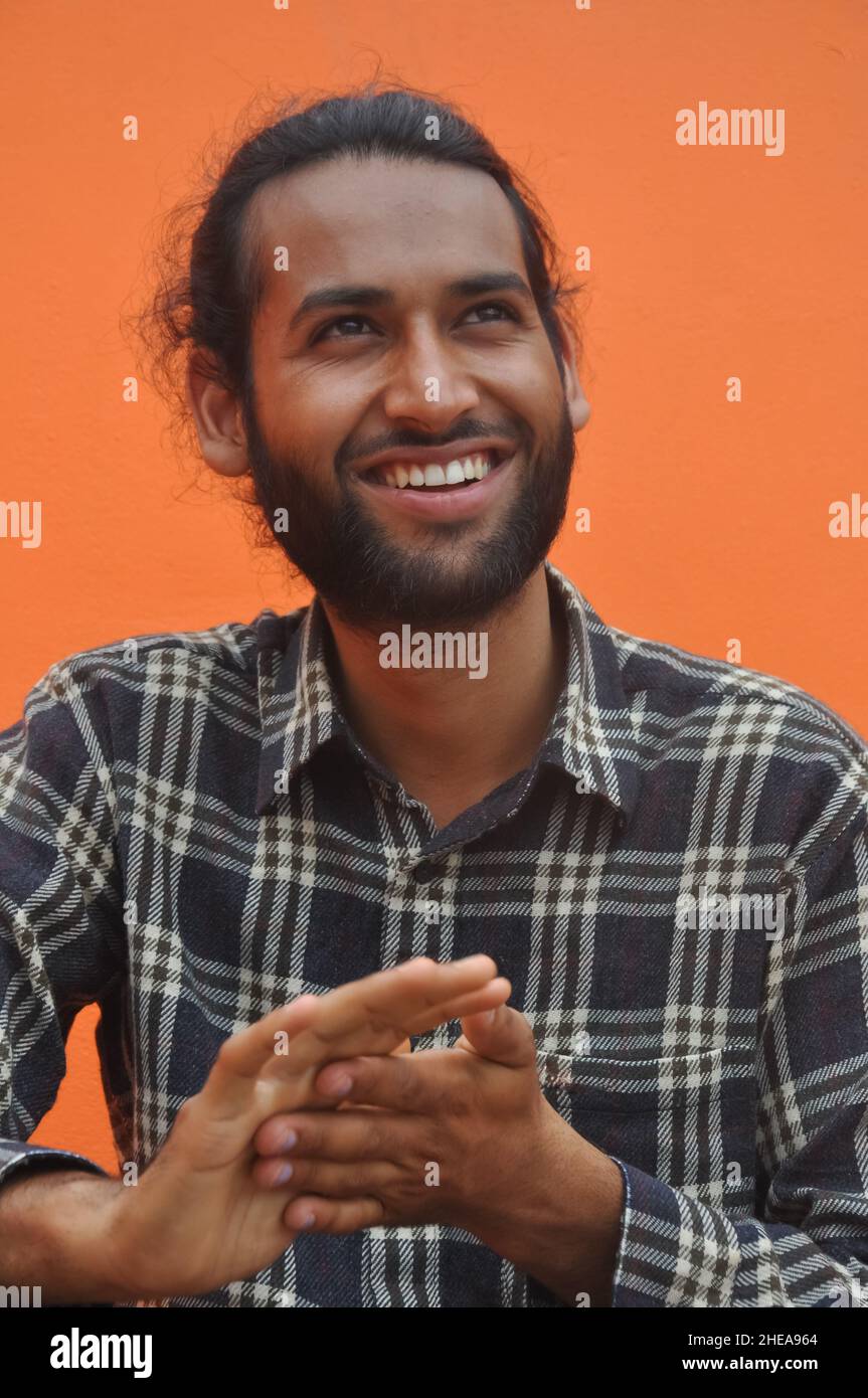Selektiver Fokus eines bärtigen jungen Mannes, der lächelt, während er seitwärts blickt und vor orangenen Wandhintergrund posiert Stockfoto