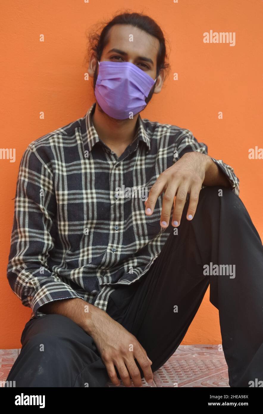 Vorderansicht eines attraktiven jungen Mannes, der eine schützende Gesichtsmaske trägt, während er draußen vor einer orangen Wand posiert und die Kamera anschaut Stockfoto