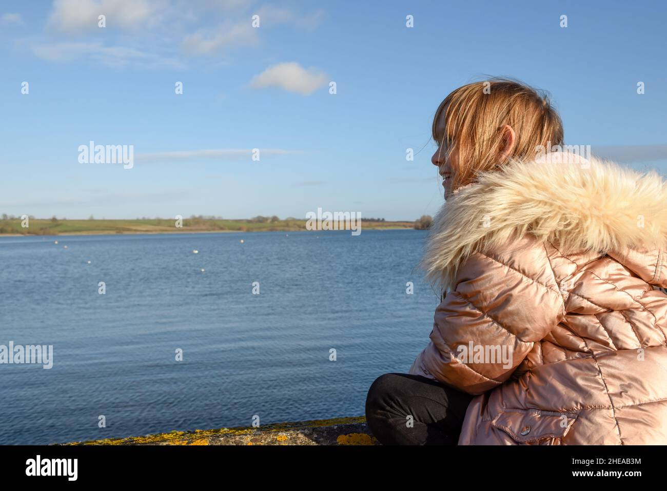 Das junge Mädchen im Mantel blickt über das Wasser und lacht an einem kalten, klaren Tag Stockfoto