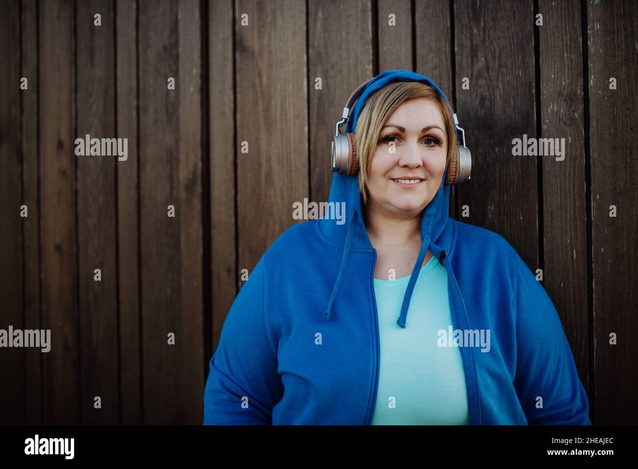 Glückliche übergewichtige Frau in Sportkleidung mit Kopfhörern und Blick auf die Kamera gegen die Holzwand Stockfoto
