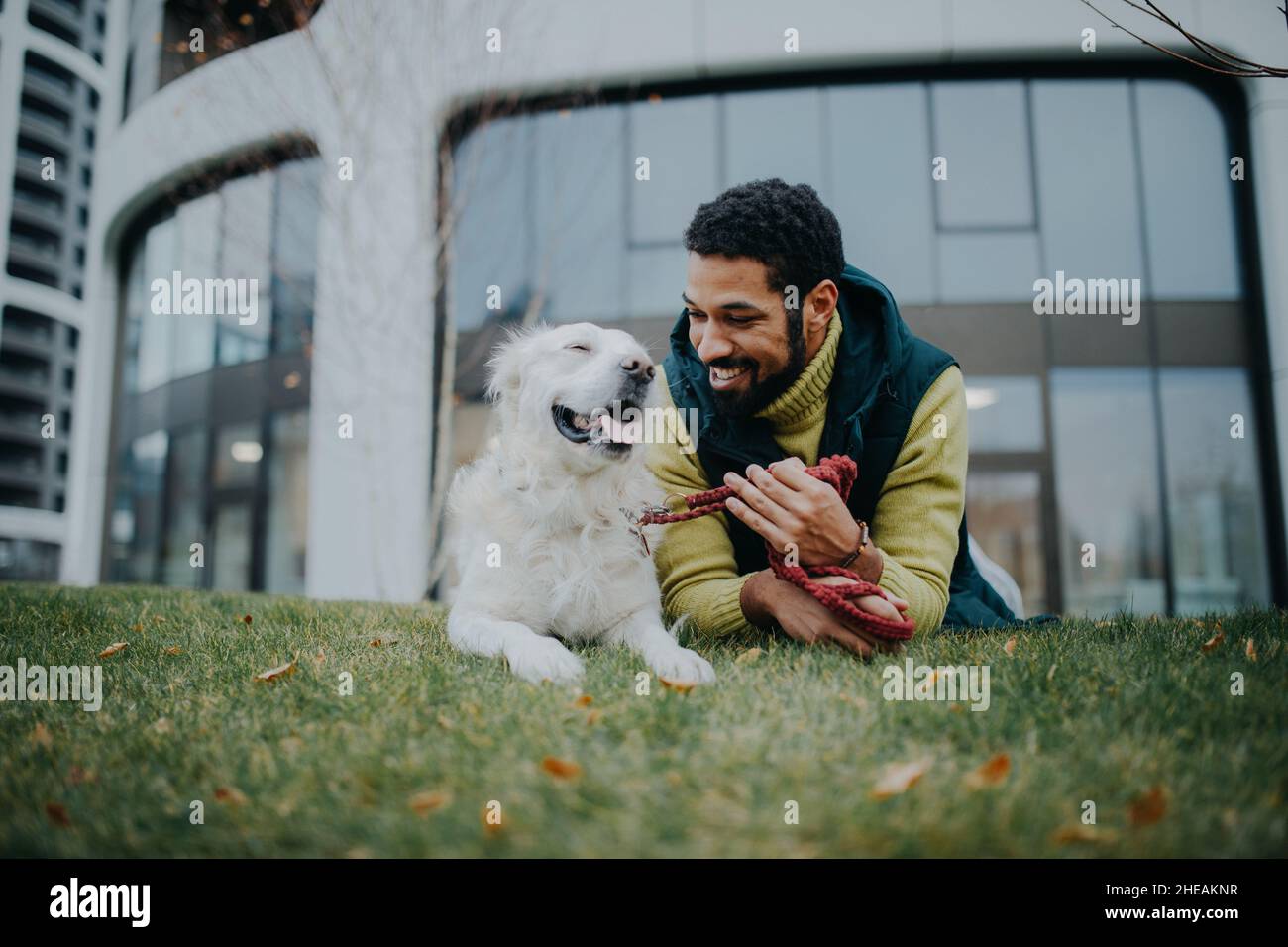 Glücklicher junger Mann, der mit seinem Hund draußen in der Stadt auf Gras liegt. Stockfoto