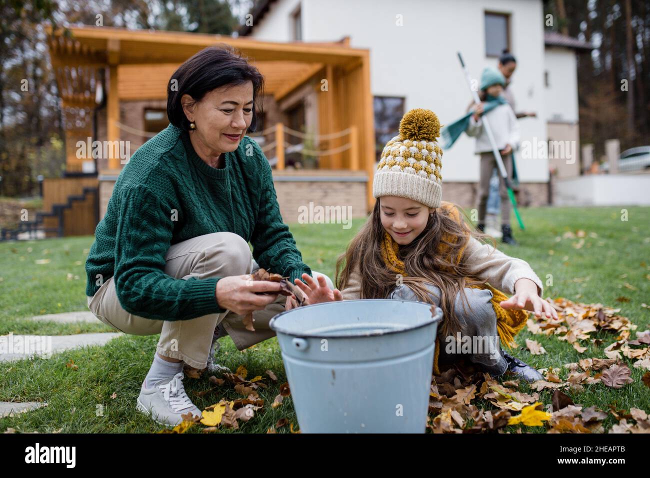 Glückliches kleines Mädchen mit Großmutter, die Blätter aufhob und sie im Herbst in den Eimer im Garten legte Stockfoto