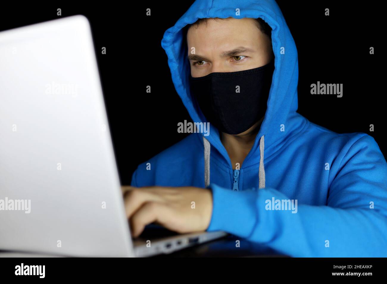 Mann in Maske und blauem Hoodie mit Laptop auf schwarzem Hintergrund. Konzept von Cyberkriminalität, Hacking und Technologie Stockfoto
