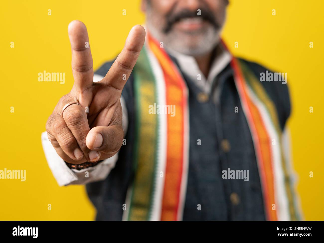 Nahaufnahme selektiver Fokus auf der Hand, nicht erkennbarer indischer Politiker, der V oder Siegeszeichen oder -Symbol zeigt, indem er die Kamera auf gelbem Hintergrund betrachtet - Stockfoto
