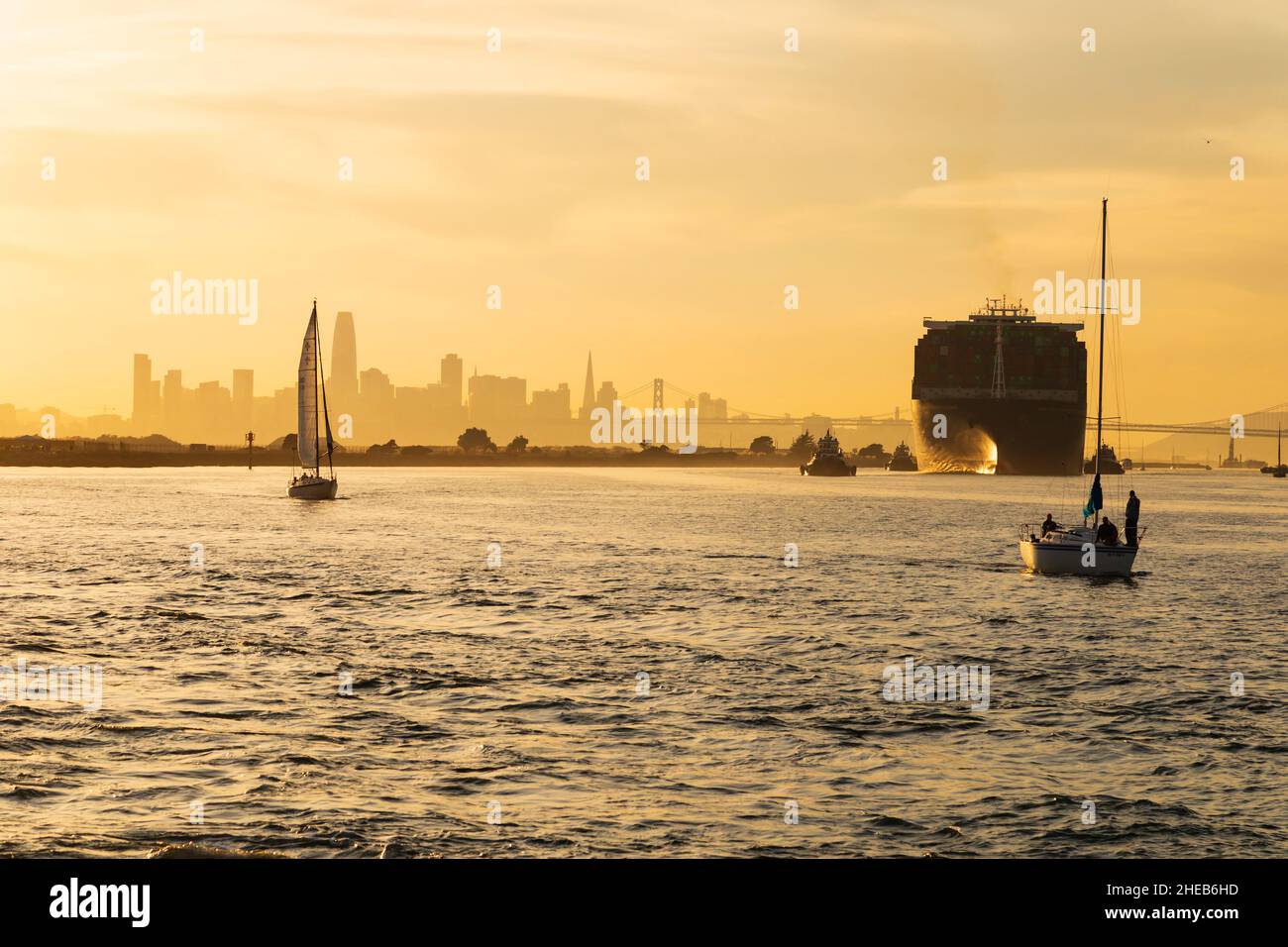 Yachten und riesige Containerschiffe fahren zum Hafen von Oakland, wenn die Sonne über der Skyline von San Francisco untergeht. Kalifornien, USA Stockfoto