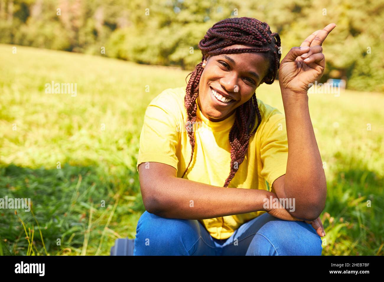 Glückliche Afrikanerin mit Rasta Zöpfen, die im Sommer in der Natur auf einer Wiese sitzt Stockfoto