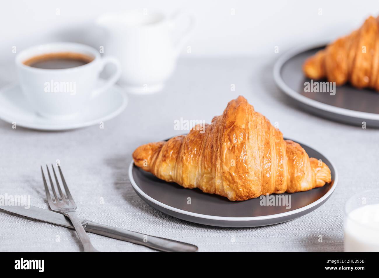 Croissant in hellweißer Umgebung, mit einer Tasse Kaffee und Milch und anderen Croissants vor verschwommenem Hintergrund. Einfache und elegante Frühstückssetting und Stockfoto