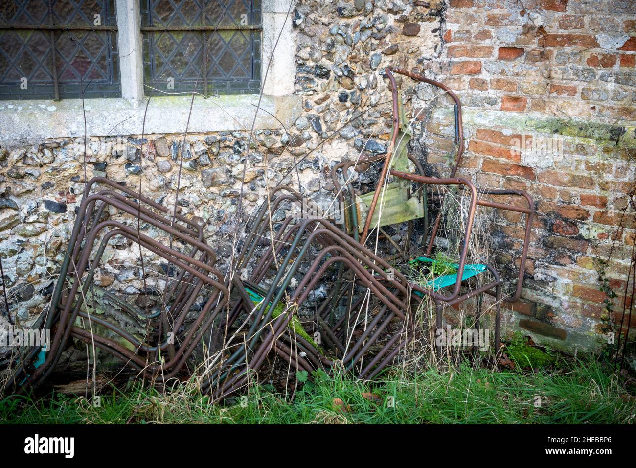 Ein kleiner Stapel rostender, metallgerahmter Stühle mit fehlender oder zerrissener Leinwand an einer Feuerstein-, Backsteinmauer und einem Pfosten-Fenster Stockfoto