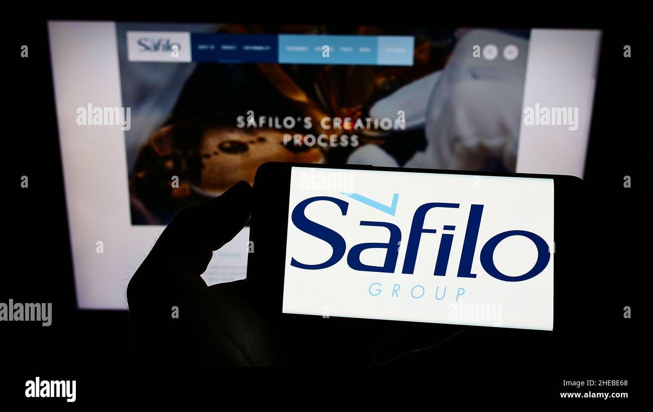 Person, die das Mobiltelefon mit dem Logo des italienischen Brillenunternehmens Safilo Group S.p.A. auf dem Bildschirm vor der Webseite hält. Konzentrieren Sie sich auf die Telefonanzeige. Stockfoto