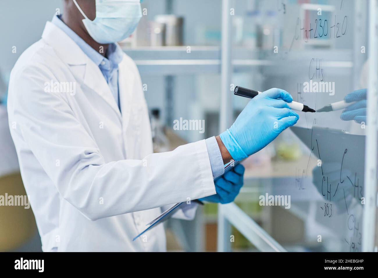 Nahaufnahme einer Chemikerin im Labormantel und Schutzhandschuhen, die während der Arbeit im Büro Formeln auf die Glaswand schreiben Stockfoto