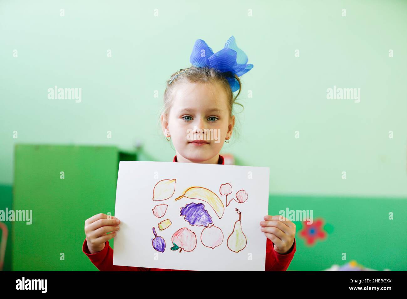 Ein Mädchen im Vorschulalter oder eine Grundschülerin hält ein Blatt Papier mit gezeichneten Früchten vor sich Stockfoto