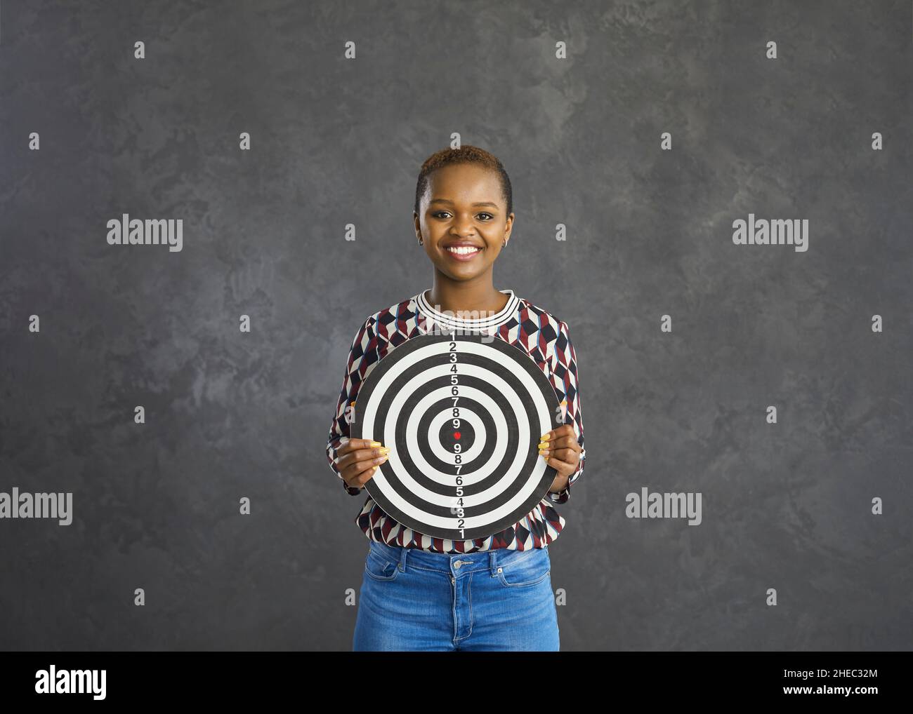 Erfolgreiche positive afroamerikanische Frau, die eine Dartscheibe auf grauem Hintergrund hält. Stockfoto