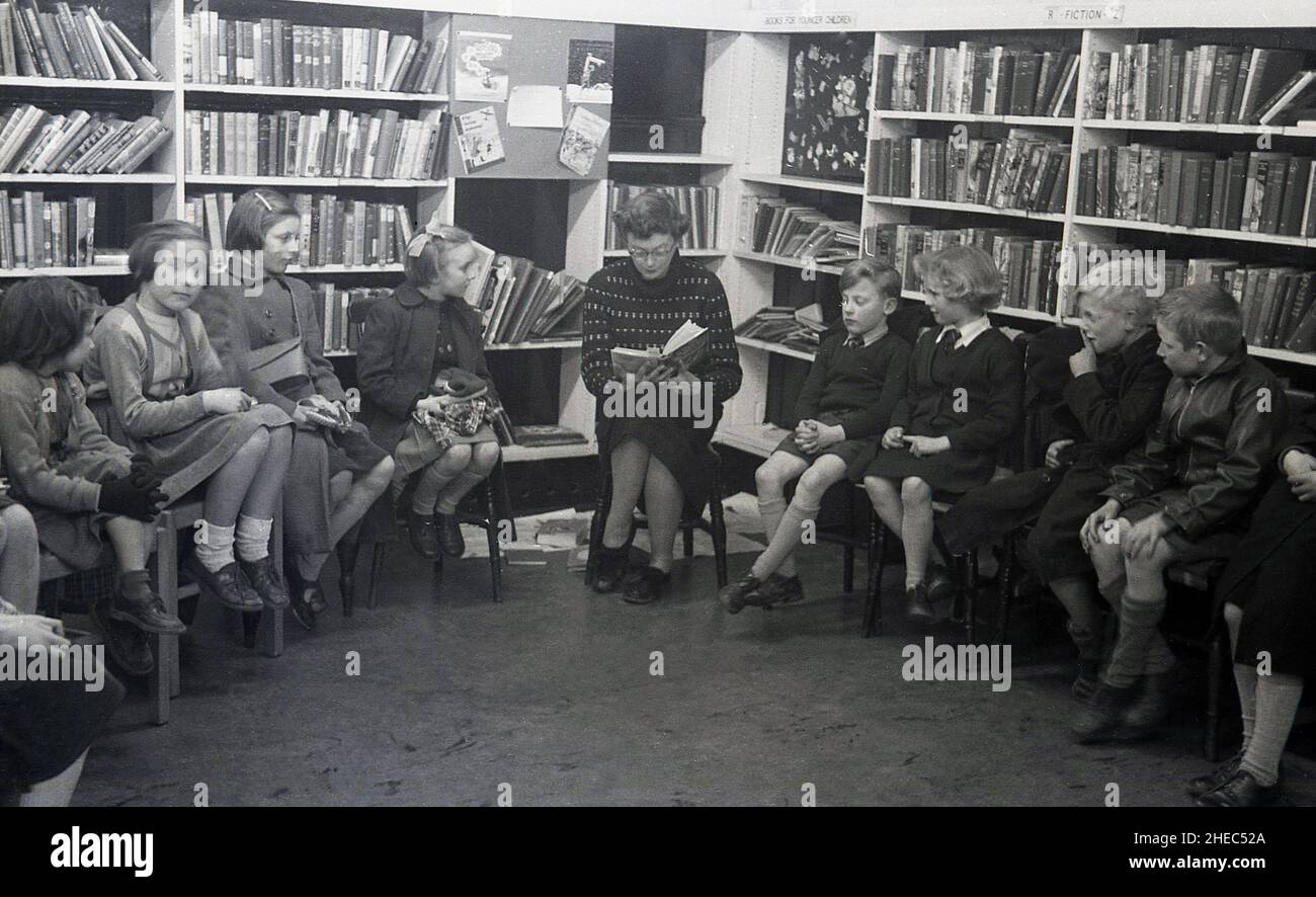1950s, historisch, in einer Schulbibliothek, liest eine Lehrerin ein Buch mit Kindern, die um sie herum sitzen und die Geschichte hören, einige tragen ihre Outdor-Mäntel, England, Großbritannien. Stockfoto